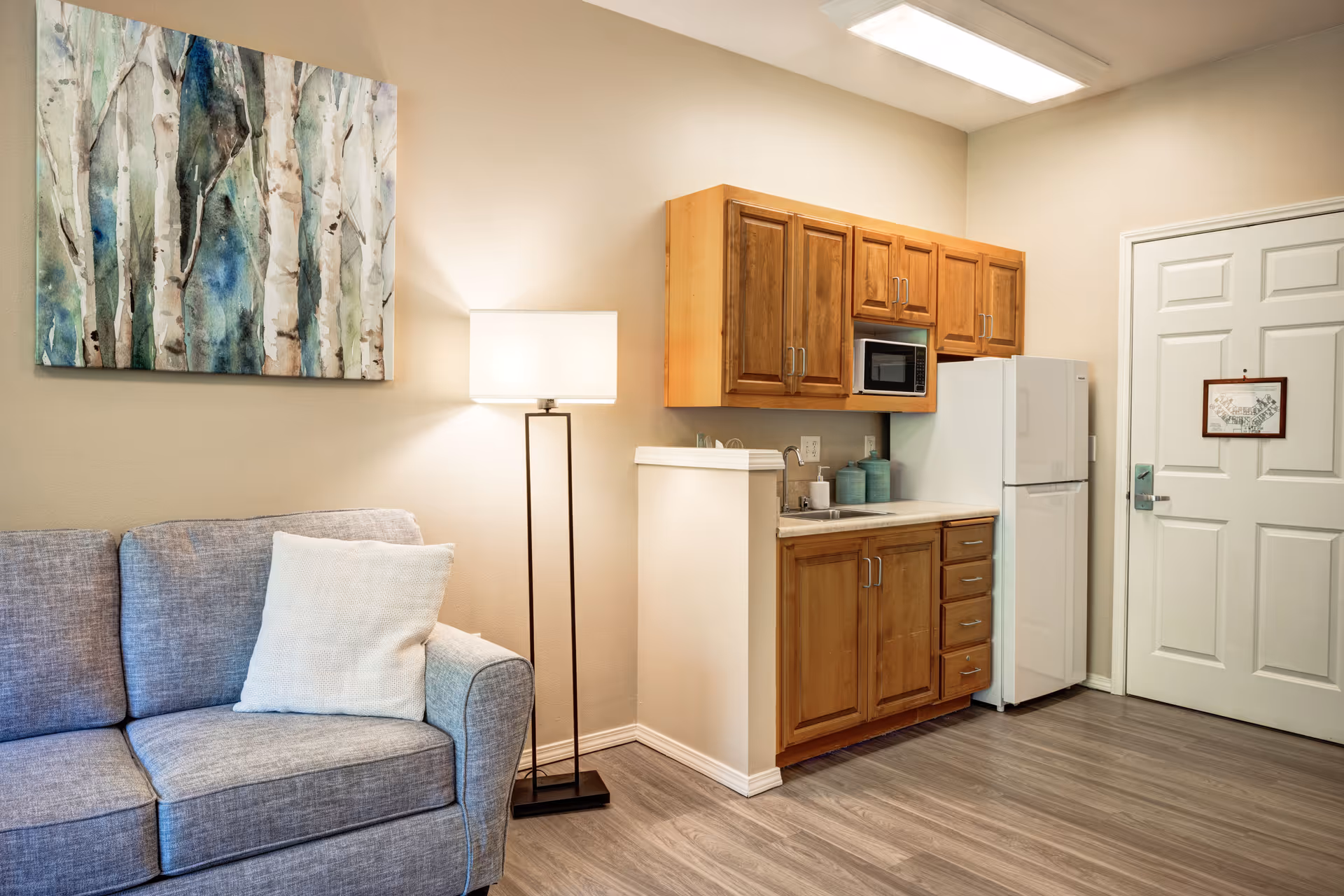 A small kitchenette area with wooden cabinets, a white refrigerator, a microwave, and a sink. Next to the kitchenette is a gray couch with a white pillow and a tall floor lamp. A painting of trees hangs on the wall above the couch. The floor is wood laminate, and there is a white door with a small framed map hanging on it.