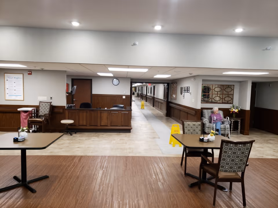 Interior view of a senior living facility hallway with a reception desk in the center background. Two tables with chairs are in the foreground on a wood-patterned floor. An elderly person is seated on a cushioned chair to the right near a side table with flowers. The hallway extends into the distance with caution signs placed along the floor.