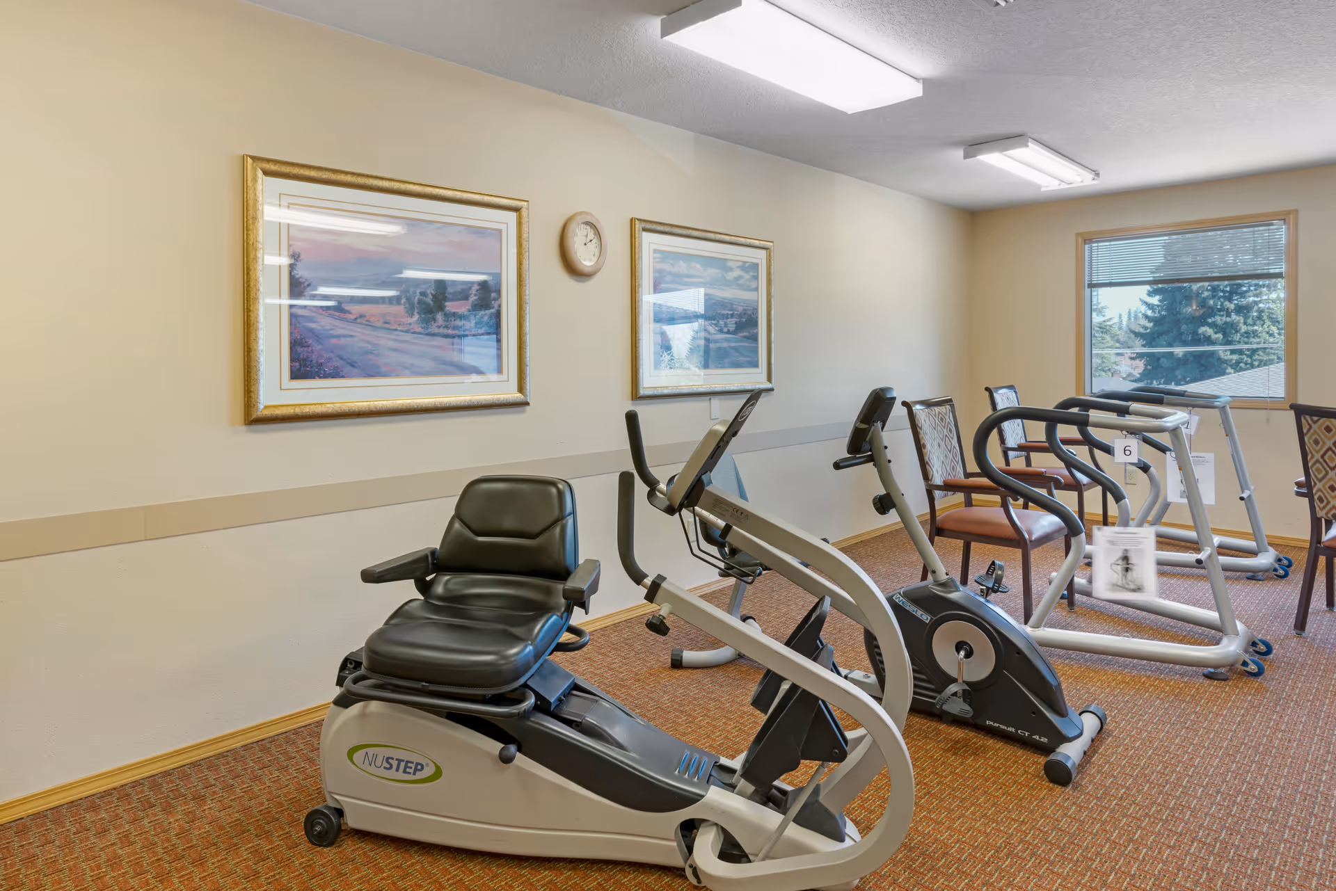A small exercise room with two stationary exercise bikes and two treadmills. The room has beige walls with two framed landscape paintings and a clock on the wall. There are also two chairs with patterned upholstery near the treadmills. A window shows trees outside.