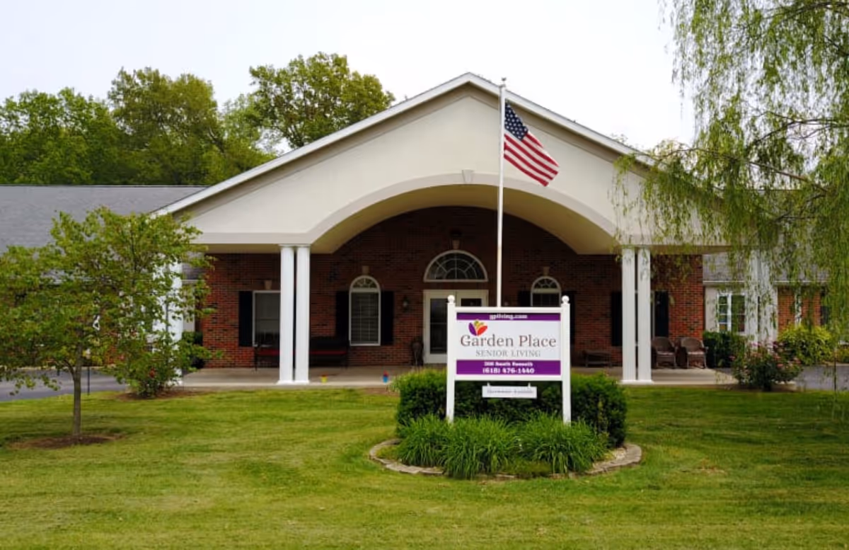Front exterior view of Garden Place Millstadt senior living facility with a brick building, white columns supporting a large archway, an American flag on a flagpole, and a sign with the facility's name and contact information surrounded by green grass and trees.