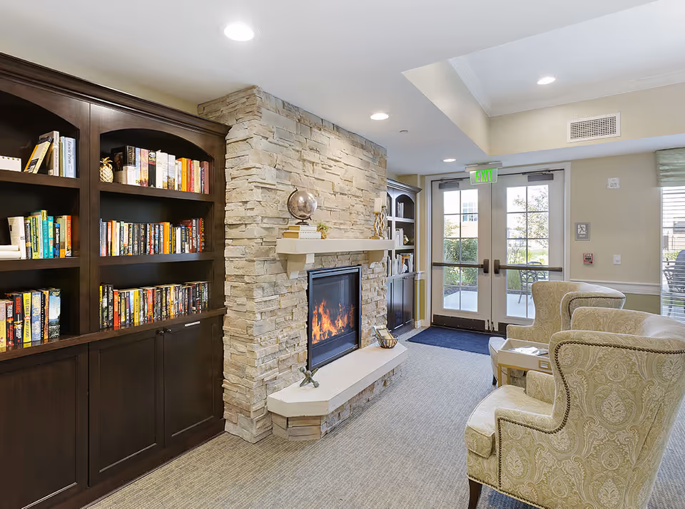 Cozy interior room with a stone fireplace, a dark wooden bookshelf filled with books, two patterned armchairs, and glass double doors leading outside.