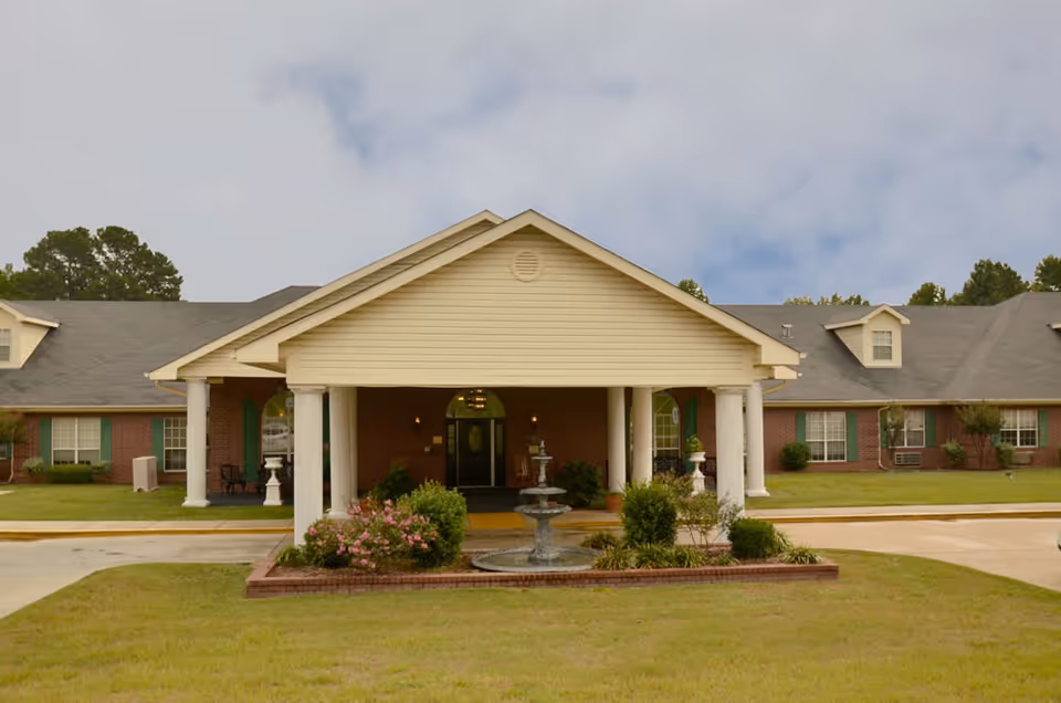 Front exterior of a single-story senior care facility with a covered portico, columns, and a fountain in front.