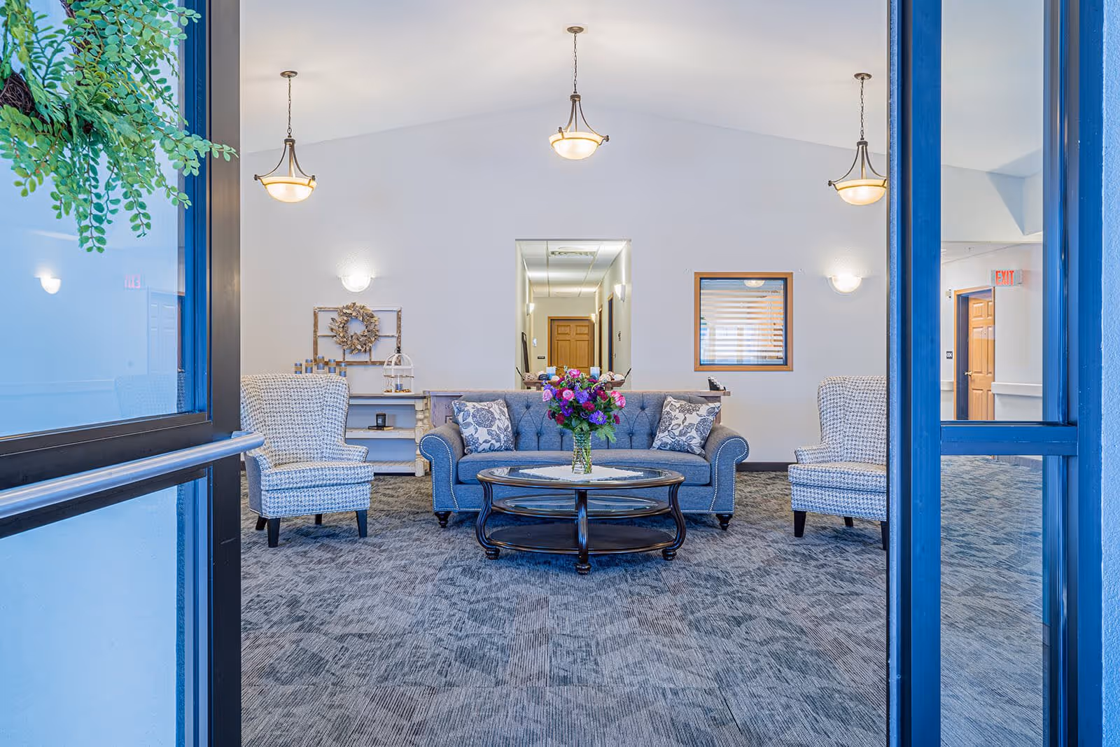 A bright and spacious living room area with a blue upholstered sofa adorned with patterned cushions, two matching armchairs, and a round wooden coffee table with a vase of colorful flowers. The room has a high vaulted ceiling with three hanging light fixtures, a carpeted floor, and a hallway visible in the background. There is a decorative wreath and shelves on the left wall and a window with blinds on the right wall.
