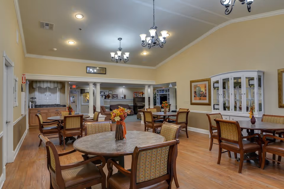 A senior living facility dining room with round tables and wooden chairs arranged neatly. Each table has a small floral centerpiece. The room features warm yellow walls, wooden flooring, chandeliers hanging from the ceiling, and framed artwork on the walls. In the background, there is a small kitchenette area and a lounge space with a piano and seating.