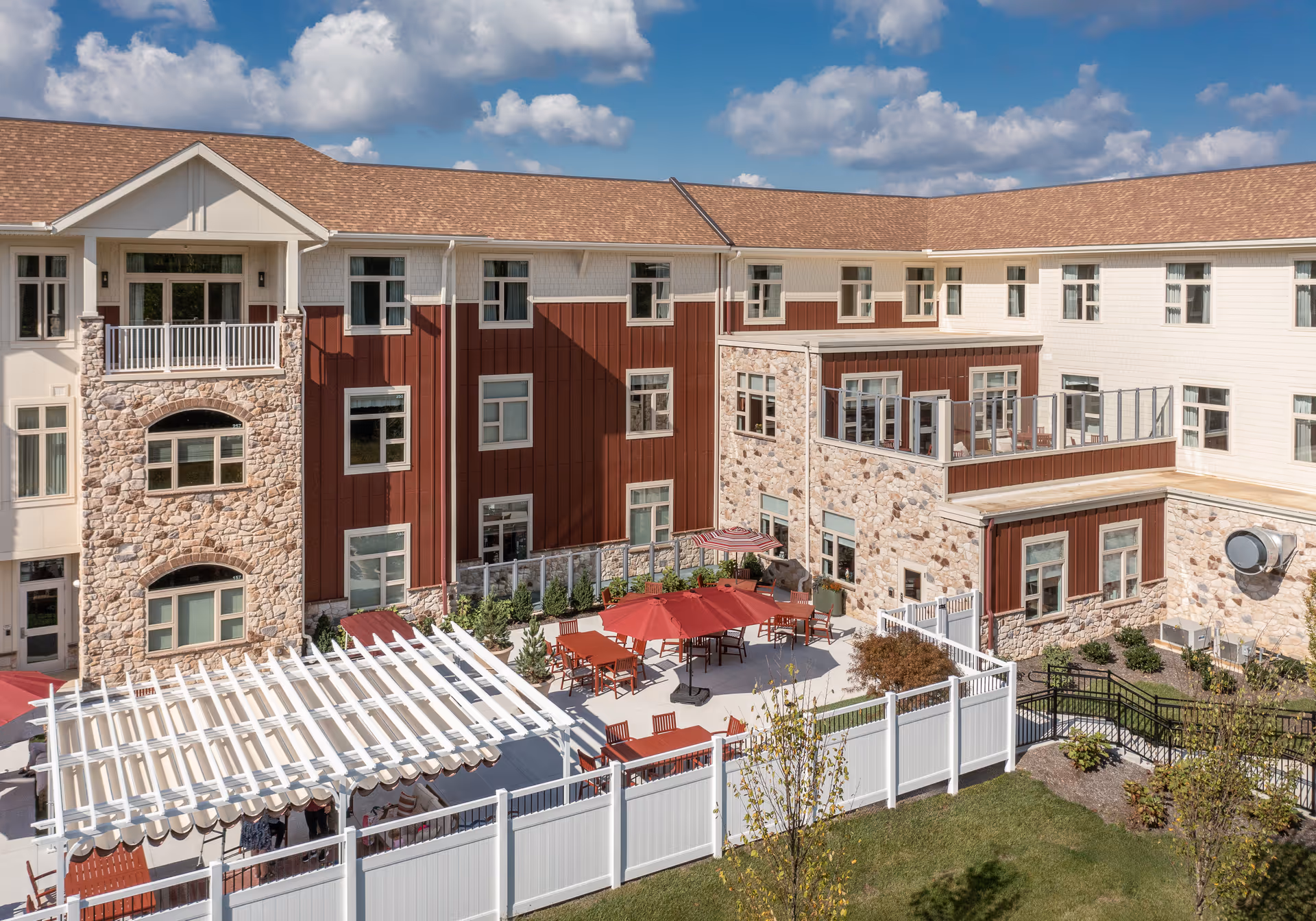 Outdoor patio area of a senior living facility with red umbrellas, tables, and chairs surrounded by a white fence. The building has a combination of stone and red siding with multiple windows and a clear blue sky with some clouds above.