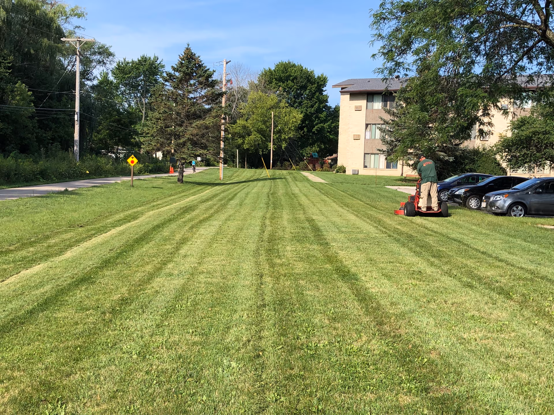 A person mowing a large, freshly cut grassy lawn next to a parking lot with several cars. In the background, there is a multi-story beige building partially obscured by trees under a clear blue sky.