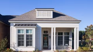 Front exterior view of a single-story house with a gray roof, white siding, and a small covered porch with two white columns. There are large windows on either side of the front door, and a clear blue sky in the background.