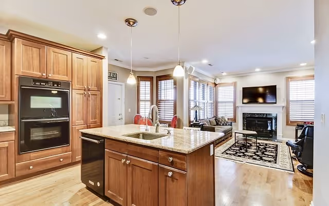 Open concept interior showing a kitchen with wooden cabinets, a granite island with a sink, and pendant lights. The kitchen opens into a living room area with a fireplace, a wall-mounted TV, a patterned rug, and seating including a sofa and chairs. Large windows with wooden blinds allow natural light to fill the space.