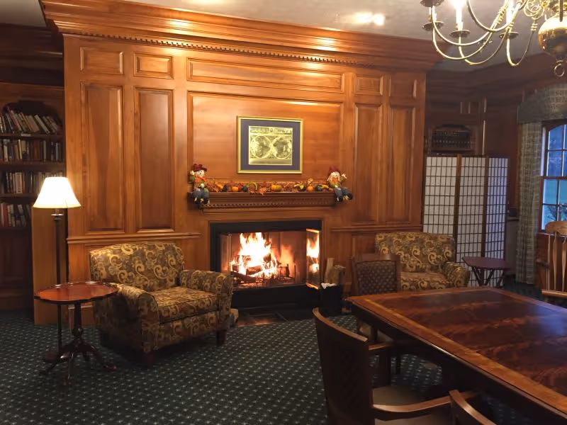 A cozy living room with a lit fireplace surrounded by wooden paneling. Two patterned armchairs flank the fireplace, with a small round table and lamp next to one chair. A large wooden table with chairs is in the foreground. Bookshelves and a folding screen are visible in the background, along with a window with curtains.