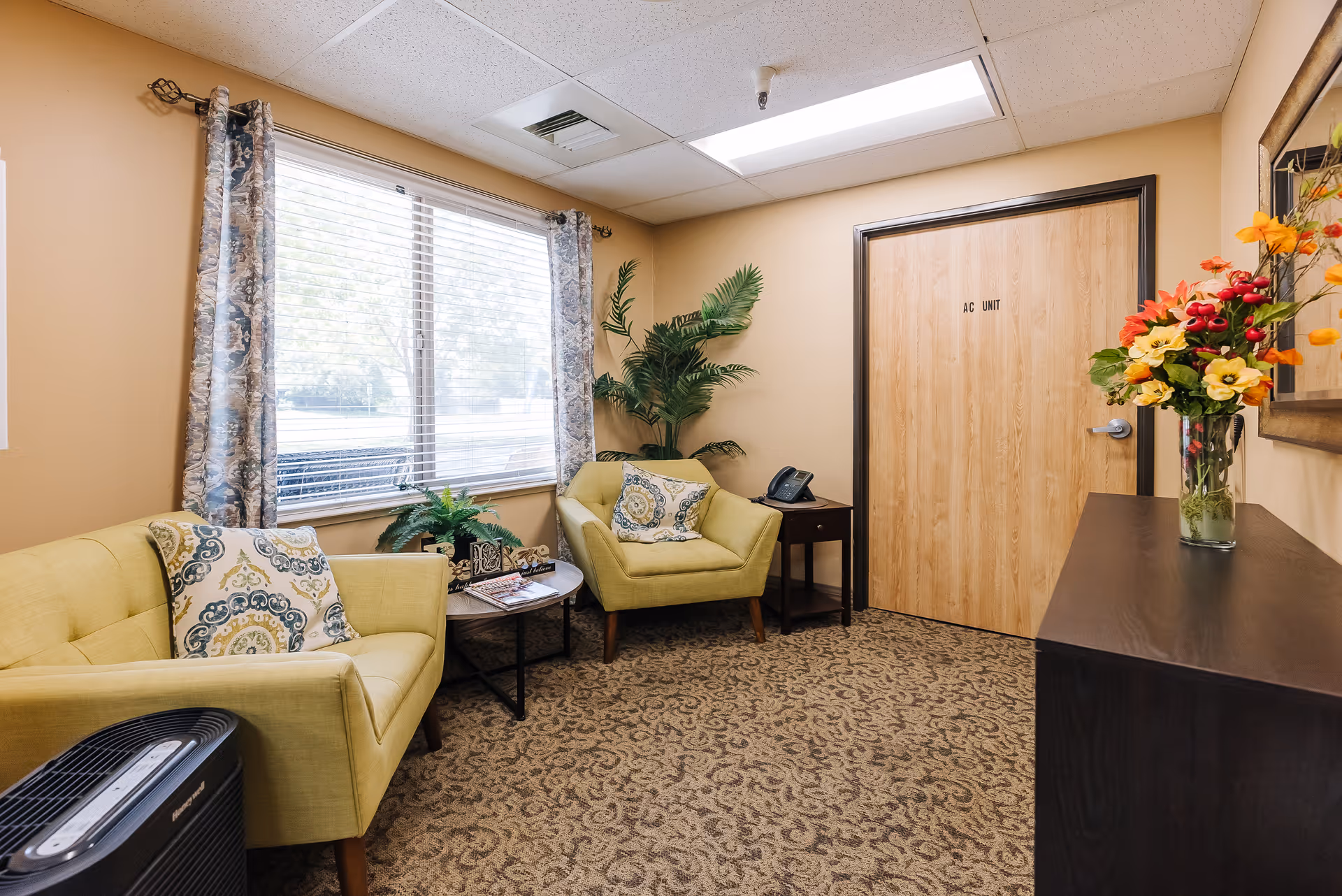 A small sitting area with two light green armchairs, each with a decorative pillow, positioned near a window with patterned curtains. There is a small round table between the chairs with magazines and a plant on it. A dark wooden cabinet with a vase of colorful flowers is on the right side, and a door labeled 'AC UNIT' is visible on the back wall. The room has beige walls and patterned carpet flooring.