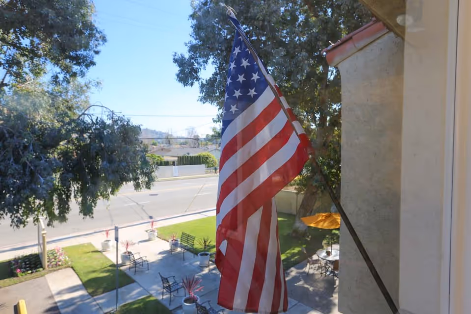 View from a window showing an American flag mounted on the exterior wall of a building. Below, there is a small outdoor seating area with benches, tables, and potted plants. Trees and a street with houses are visible in the background under a clear blue sky.
