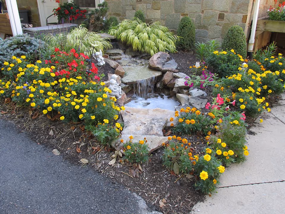 A small landscaped garden area with a stone water feature that has a gentle waterfall flowing into a small pool. The garden is bordered by vibrant yellow and red flowers, green shrubs, and decorative stone statues of children. The background shows a stone wall and part of a building entrance.