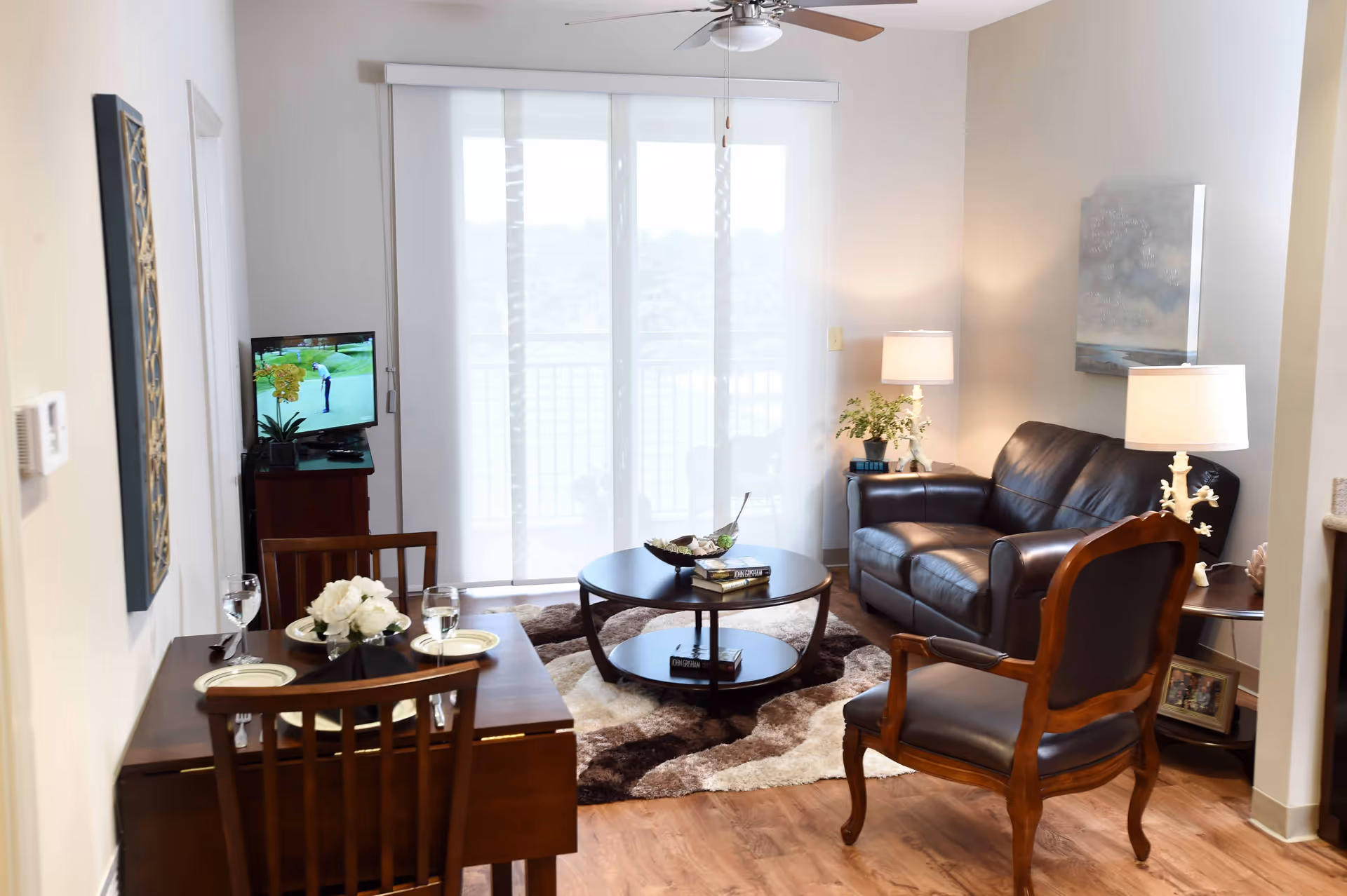 A cozy living room and dining area with wooden flooring. The living room features a dark leather sofa, a wooden armchair with leather cushions, a round coffee table on a patterned rug, and two side tables with lamps and plants. The dining area has a small wooden table set for two with plates, glasses, and a flower centerpiece. A TV is placed on a stand near sliding glass doors with sheer curtains, allowing natural light to fill the room.