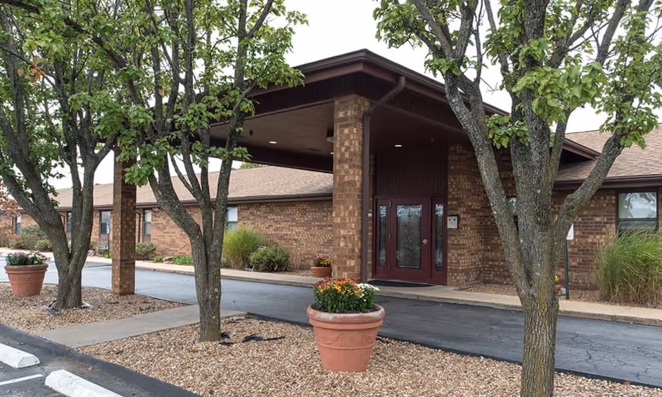 Brick nursing center entrance with a covered porte-cochère, double glass doors, trees and potted planters.