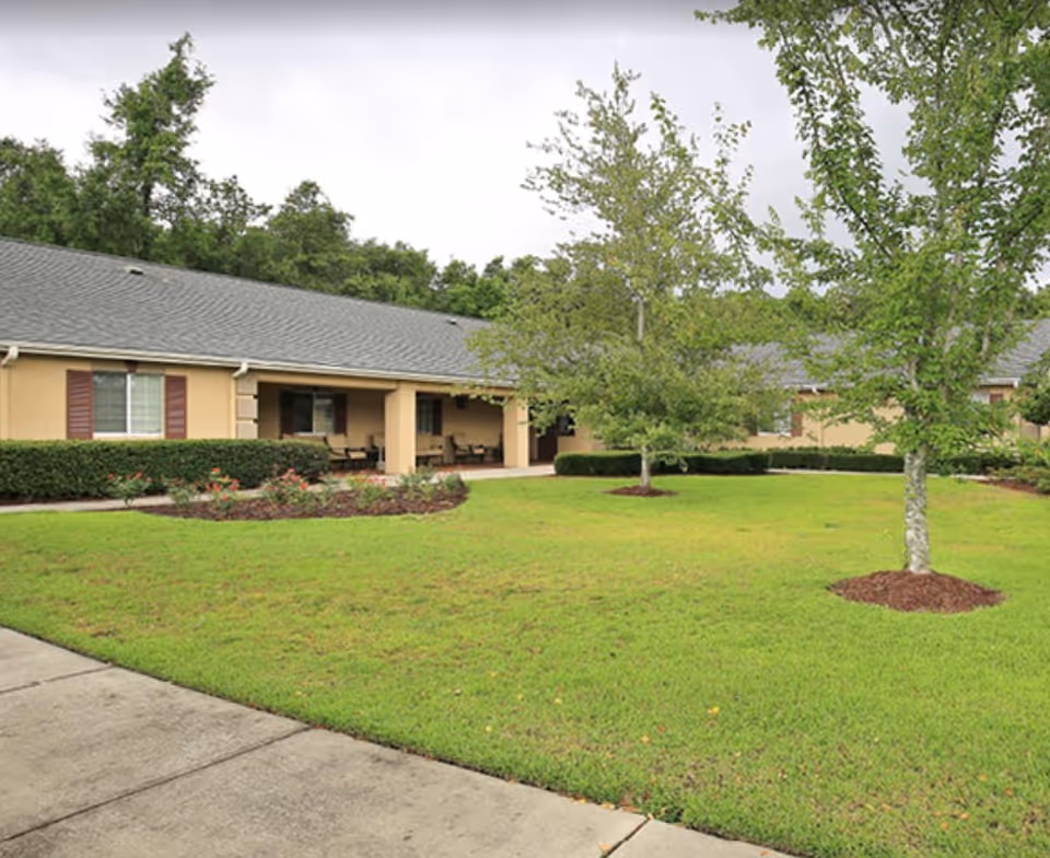 Single-story beige senior living building with a covered porch facing a landscaped lawn with trees and a sidewalk.