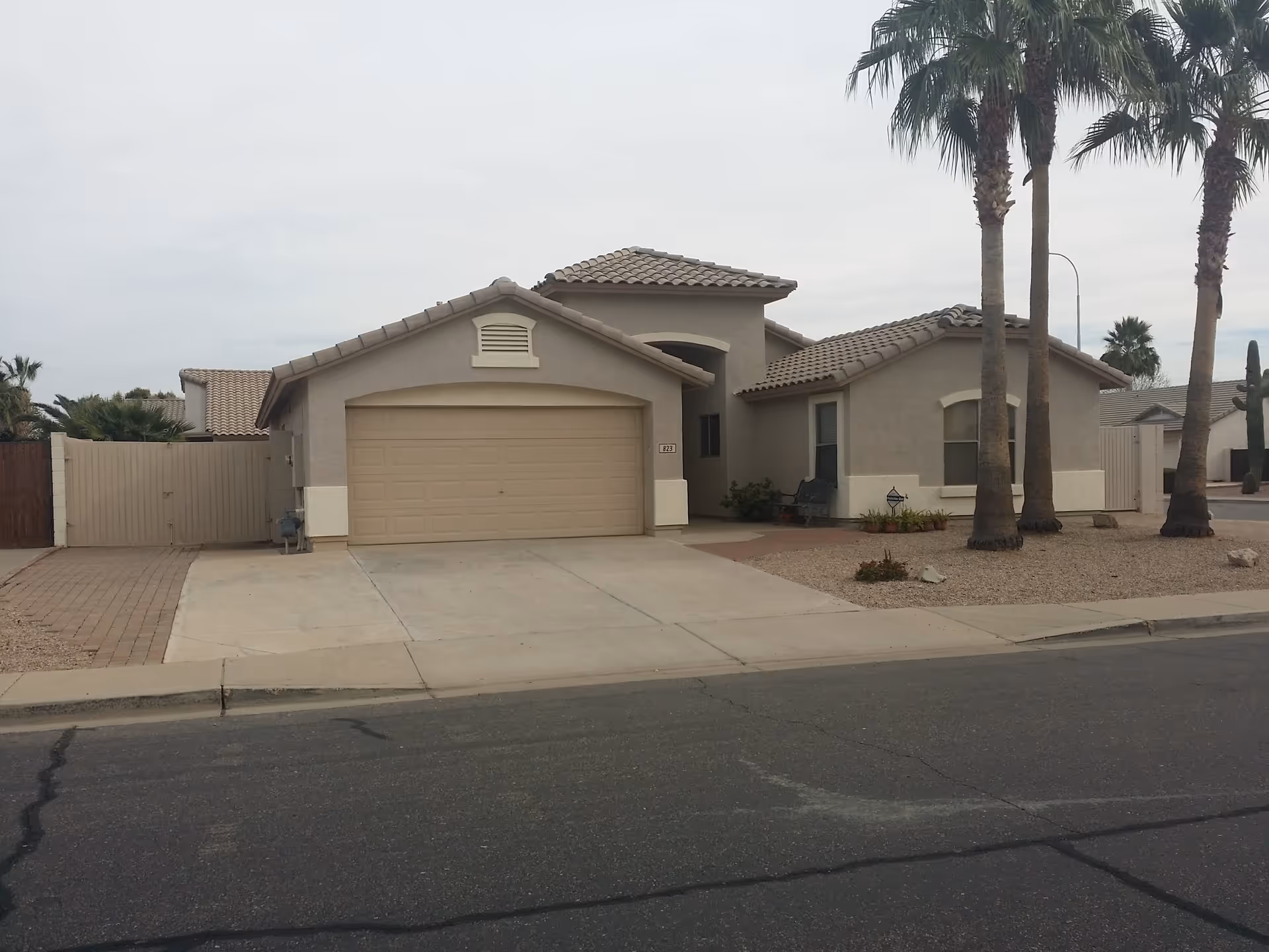 Front exterior view of a single-story house with a two-car garage, beige stucco walls, tiled roof, and palm trees in the front yard. The driveway and sidewalk are visible, along with desert landscaping featuring gravel and small plants.