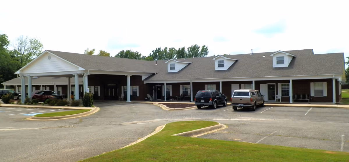 Exterior view of Clarksville Retirement Center showing a single-story building with a covered entrance supported by white columns, several windows, and a parking lot with a few parked vehicles in front.