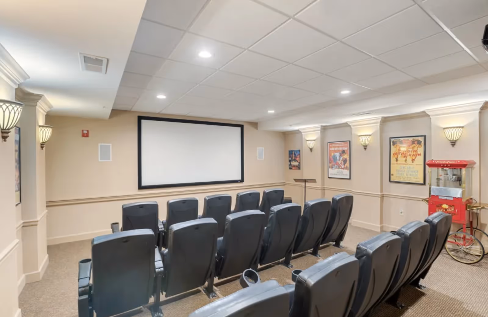 A small movie theater room with two rows of black theater seats facing a large blank projection screen. The room has beige walls with decorative molding, wall sconces providing soft lighting, framed vintage movie posters on the walls, and a red popcorn machine on the right side.