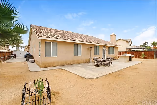 Backyard view of a single-story house with a tiled roof, beige exterior walls, and several windows. The yard has a concrete patio area with an outdoor dining table and chairs, a barbecue grill, and a small fenced plant in the dirt yard. A wooden fence encloses the yard, and neighboring houses are visible in the background under a clear blue sky.