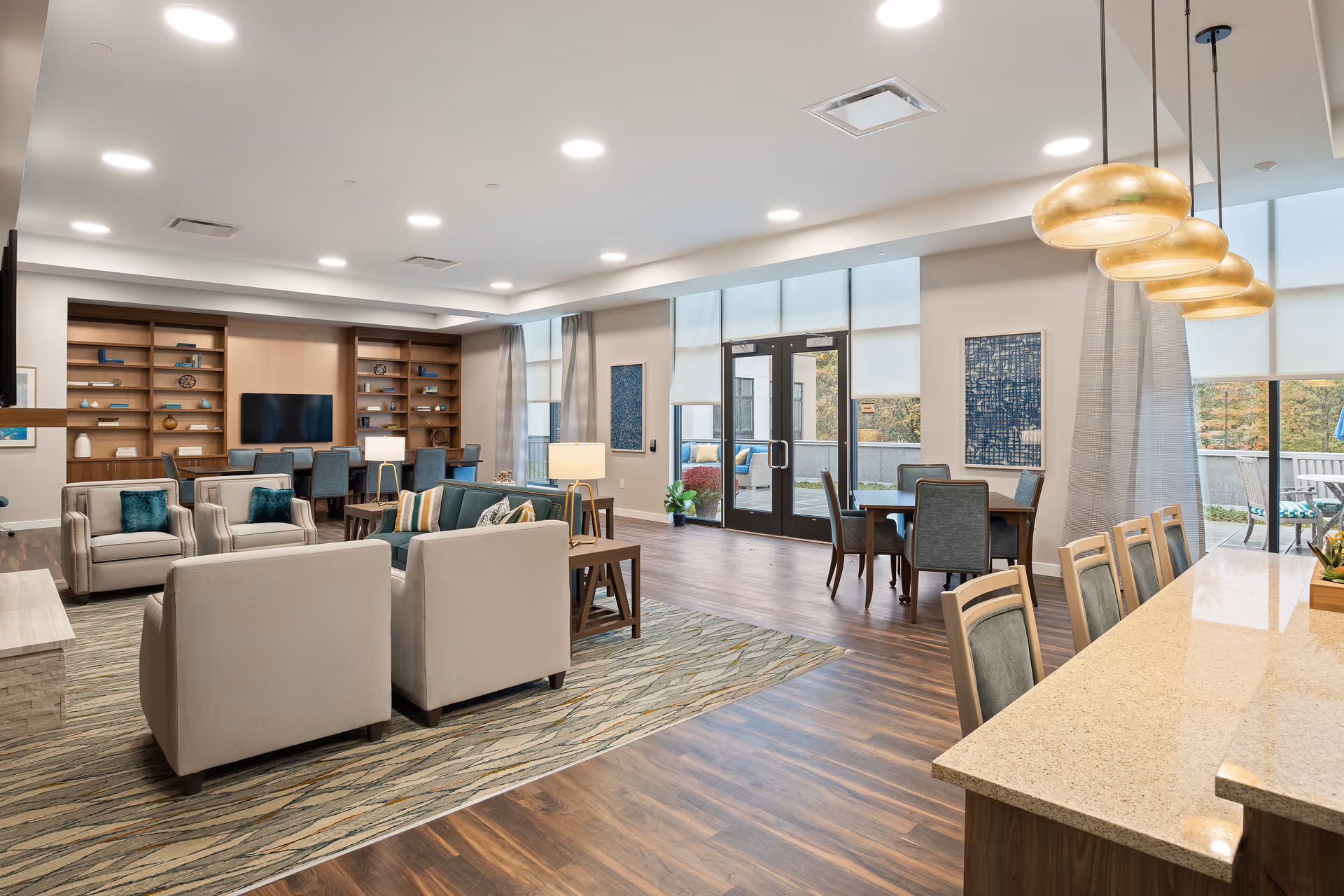 A spacious and well-lit senior living common area featuring comfortable seating with armchairs and sofas arranged around a rug, a large wooden bookshelf with decorative items and a TV, multiple dining tables with chairs, large windows with roller shades, and a kitchen counter with pendant lights hanging above.