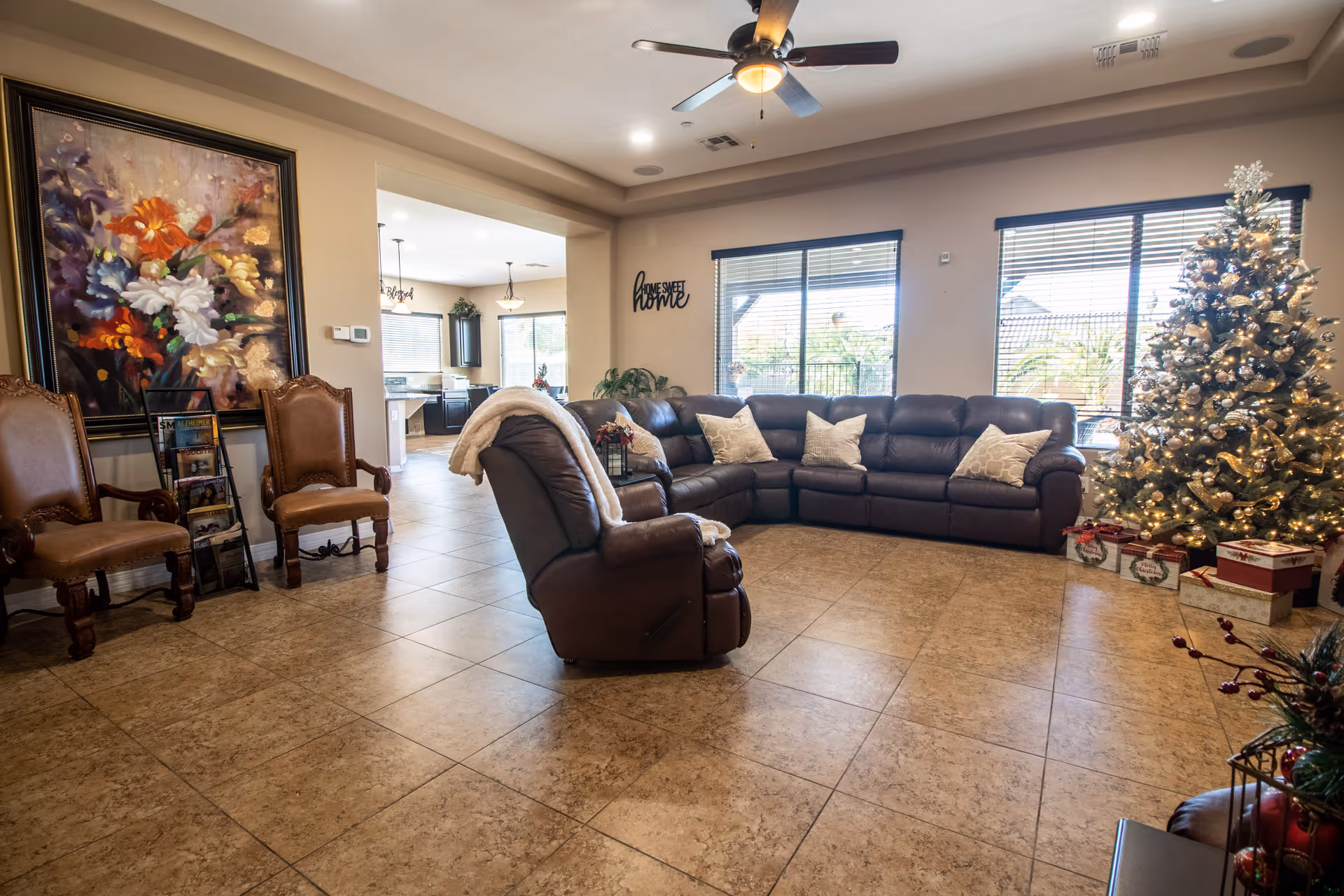 A spacious living room with a large brown leather sectional sofa adorned with beige pillows, a matching recliner with a white throw blanket, and two wooden chairs with leather seats. A decorated Christmas tree with lights and wrapped presents is placed near large windows with blinds. The room has tiled flooring, a ceiling fan with a light, a large colorful floral painting on the wall, and a magazine rack. The adjacent kitchen area is visible through an open doorway.
