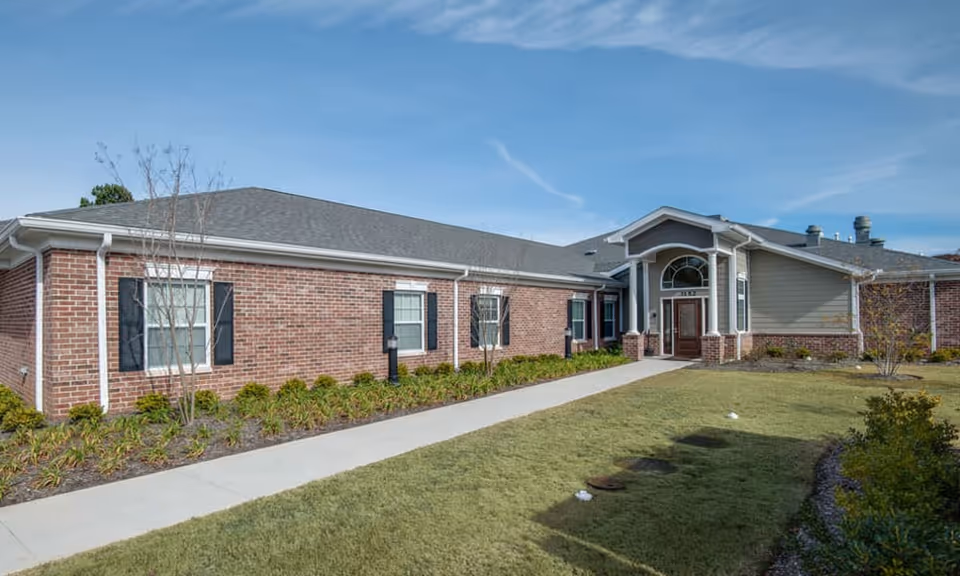Exterior view of a single-story senior living facility building with brick and siding walls, several windows with black shutters, a paved walkway leading to the main entrance, and a well-maintained lawn and landscaping under a clear blue sky.