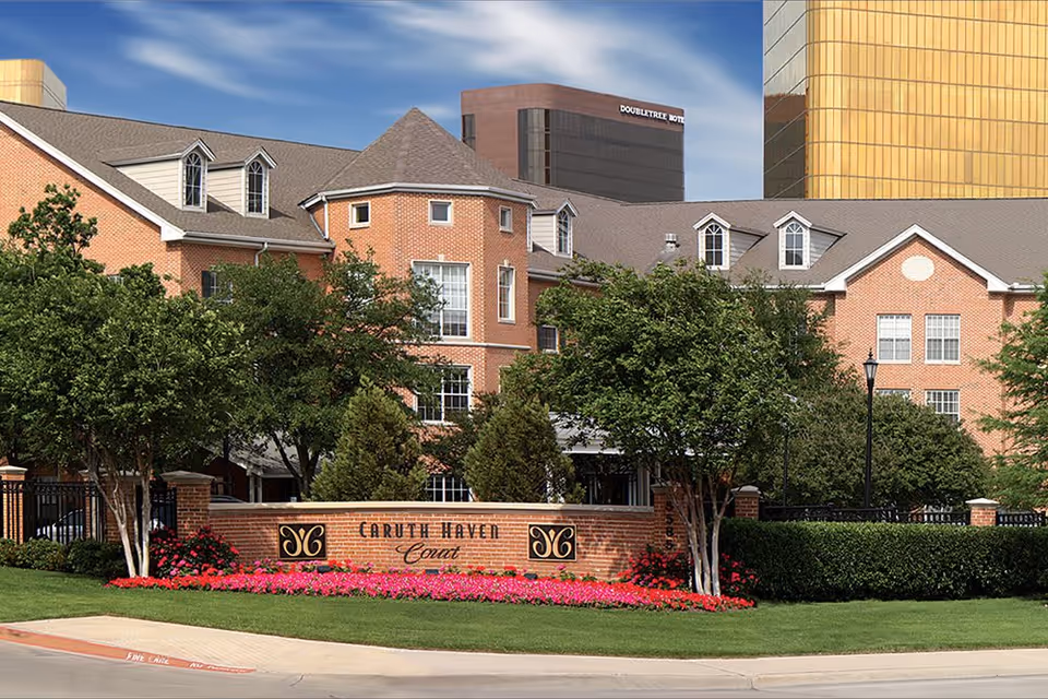 Exterior view of Caruth Haven Court, a brick building with multiple windows and dormers, surrounded by trees and a well-maintained lawn with pink flowers in front. A brick sign with the facility name is visible near the entrance. In the background, taller buildings including one labeled DoubleTree Hotel can be seen under a partly cloudy sky.