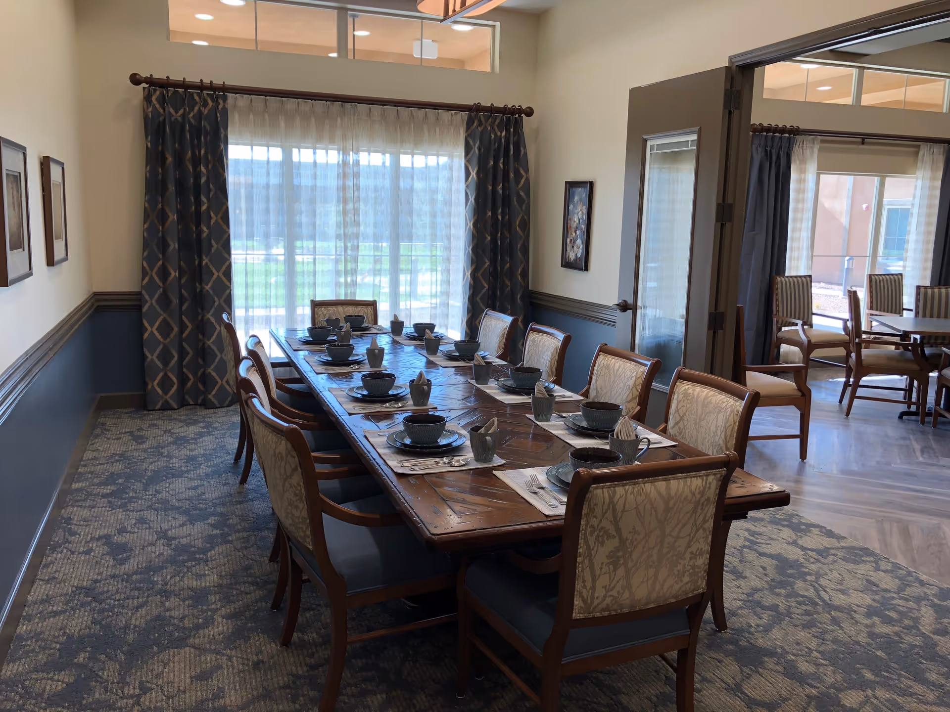 A long wooden dining table set with place settings and upholstered chairs in a formal dining room with large windows and curtains.
