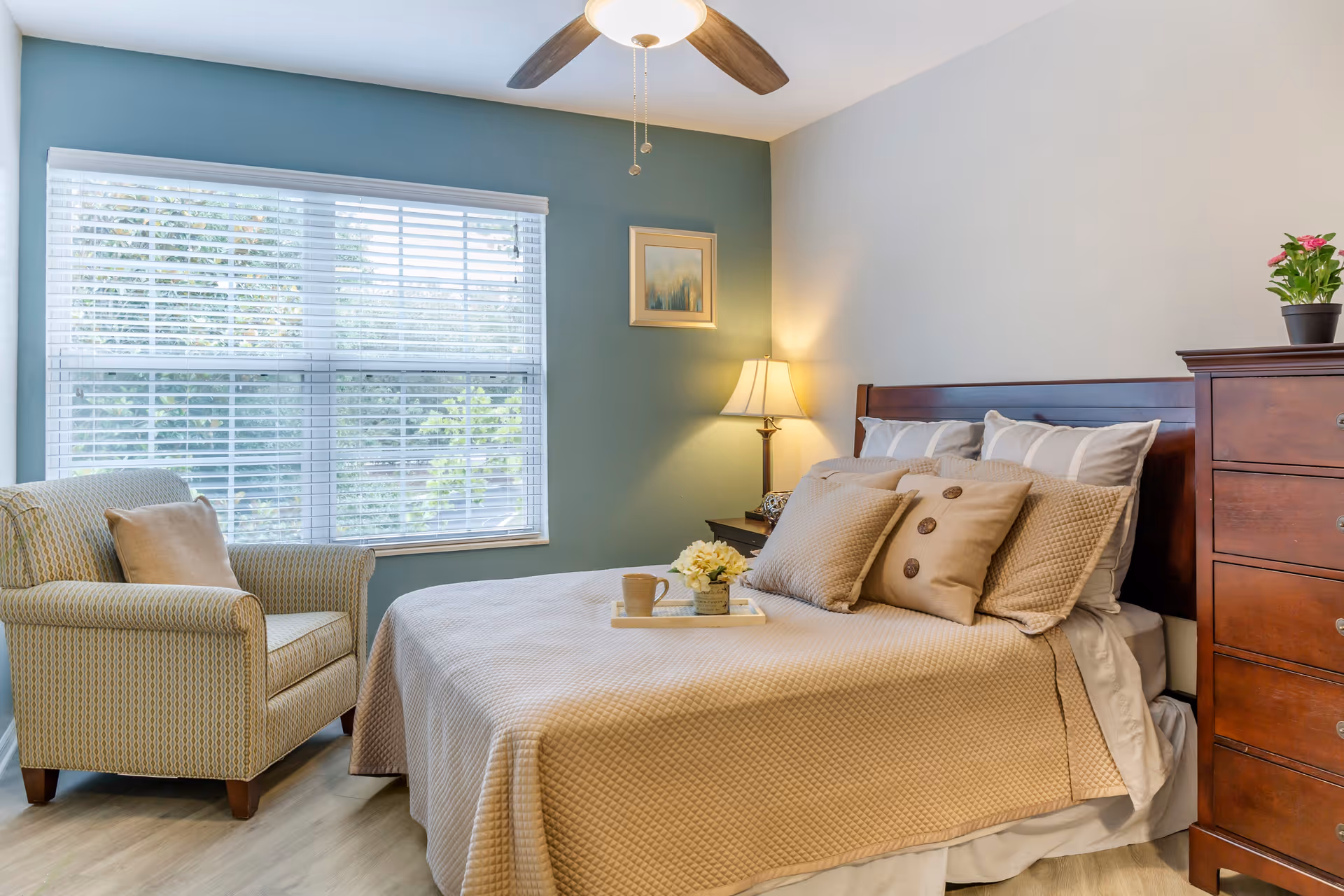 A cozy bedroom with a neatly made bed featuring beige bedding and multiple pillows. A wooden headboard and matching dresser are on the right side. A cushioned armchair with a pillow is positioned near a large window with white blinds. A small table lamp is lit on a nightstand beside the bed, and a ceiling fan with a light fixture is above.
