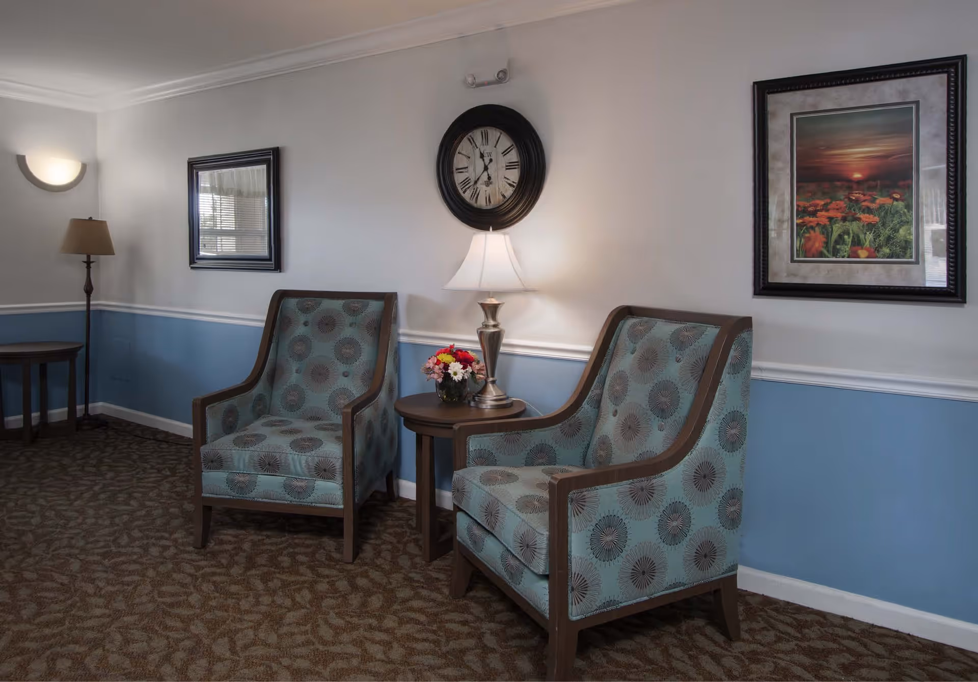 A cozy seating area with two patterned armchairs separated by a small round wooden table holding a lamp and a small flower arrangement. The walls are painted white on the upper half and blue on the lower half, with a chair rail molding in between. A large round clock hangs on the wall above the table, and two framed pictures are visible on the walls. The floor is carpeted with a patterned design.