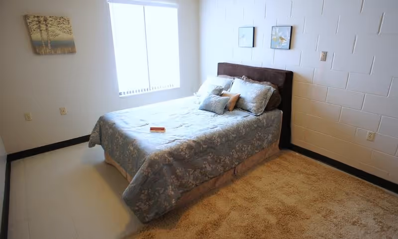 A simple bedroom with a bed covered in a blue floral bedspread and several pillows. The room has a window with blinds letting in natural light, a beige carpet on part of the floor, and white painted brick walls with two small framed pictures hanging above the bed. There is also a painting on the adjacent wall.