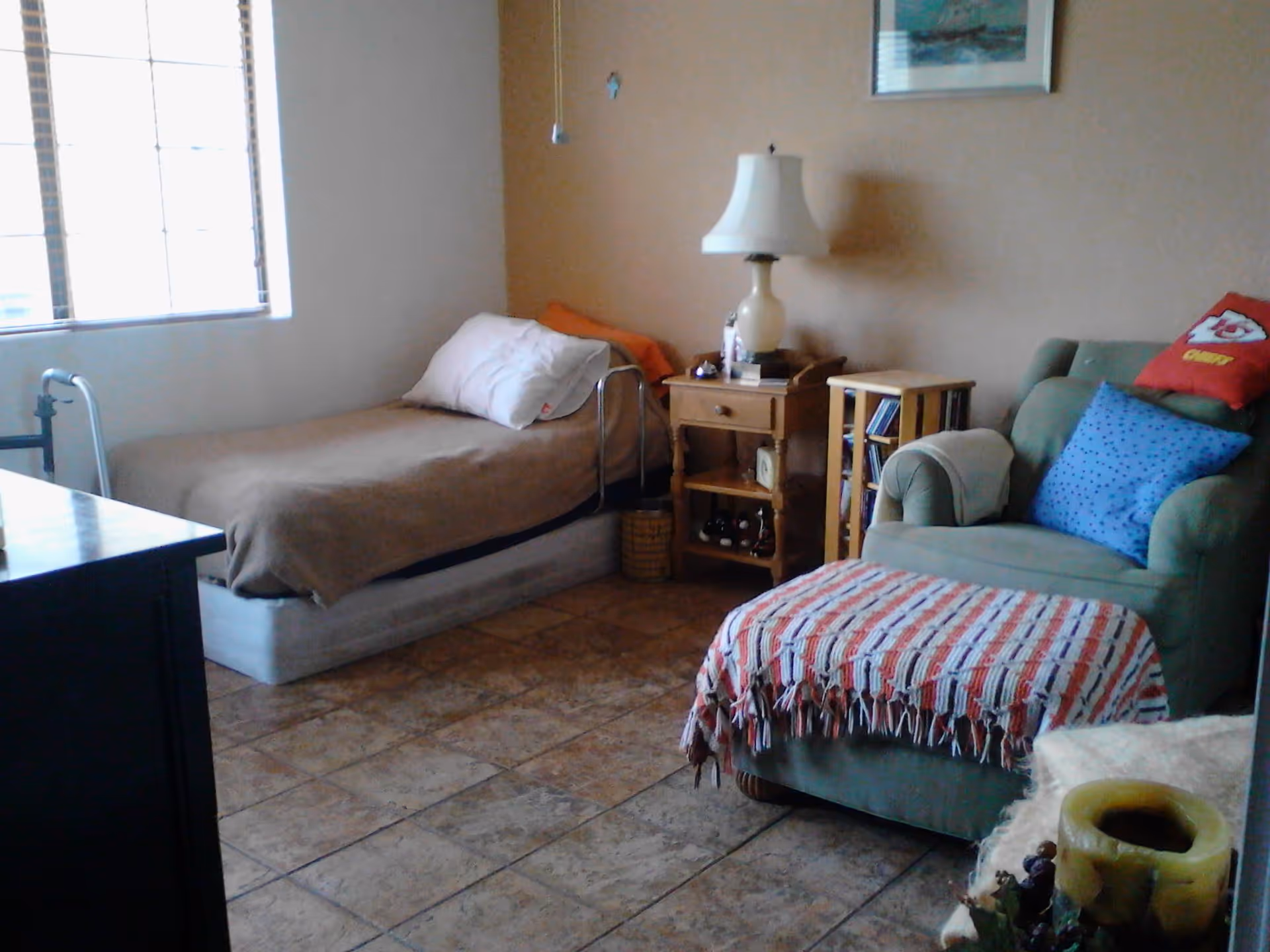 A cozy bedroom with a single bed covered with a brown blanket and a white pillow near a window with blinds. Next to the bed is a wooden nightstand with a lamp, books, and small decorative items. There is a green armchair with a blue pillow and a red pillow with a Kansas City Chiefs logo. A small wooden shelf with CDs is beside the nightstand. The floor is tiled, and a colorful woven blanket is draped over the armchair's ottoman.
