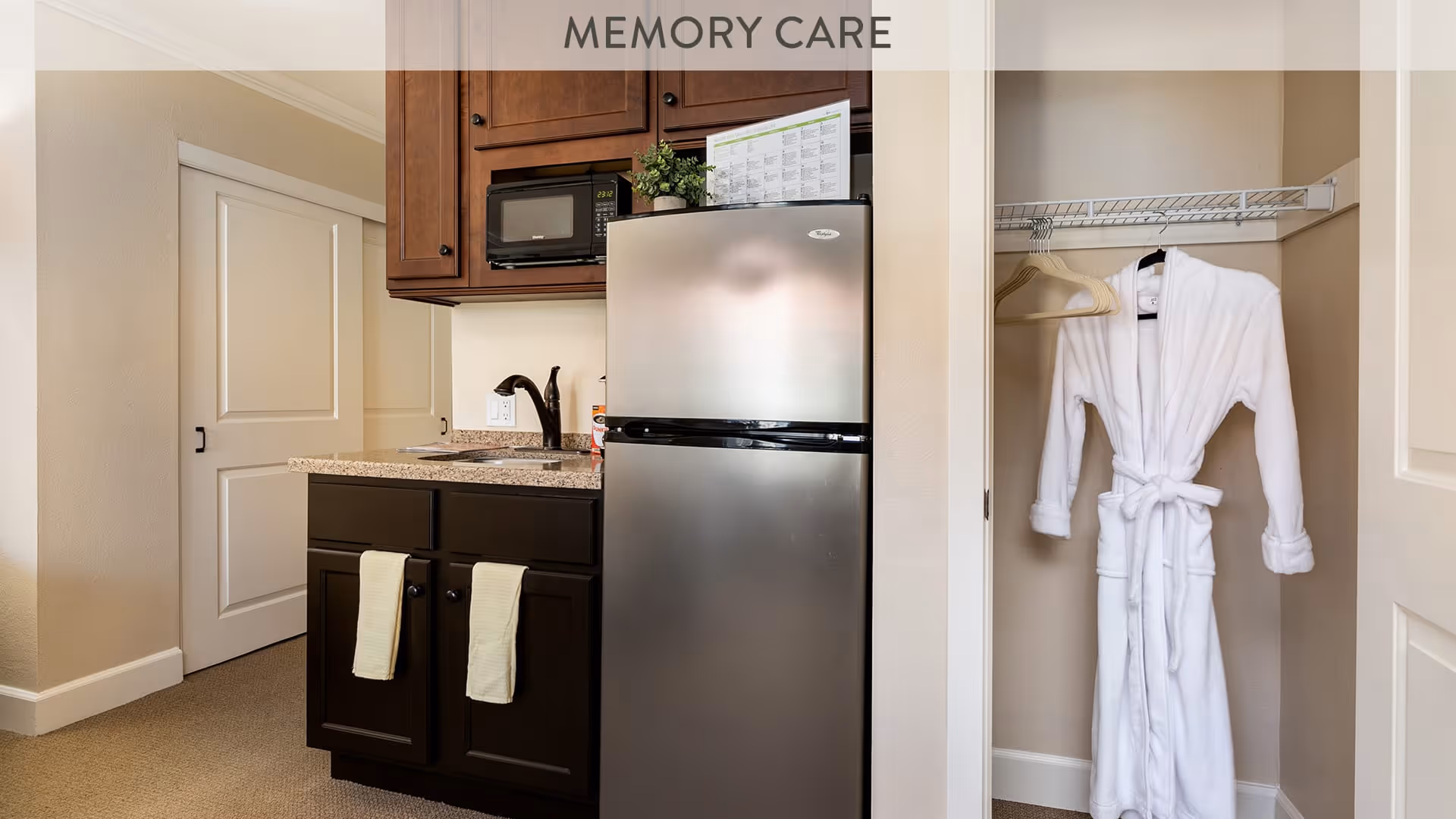 A small kitchenette area with a stainless steel refrigerator, dark wood cabinets, a microwave, and a granite countertop with a sink. Two light-colored towels hang on the cabinet doors below the sink. To the right, there is an open closet with a white bathrobe hanging on a hanger. The walls are painted beige, and there is a sliding door to the left.