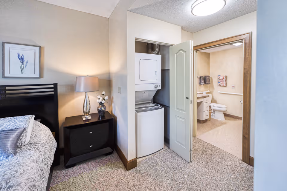 A bedroom corner featuring a bed with patterned bedding and pillows, a dark wooden nightstand with a lamp and a vase of flowers, a closet with a stacked washer and dryer, and an open door leading to a bathroom with a toilet, sink, and grab bars.