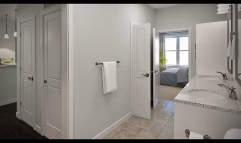 Interior view of a senior living facility bathroom area with a double sink vanity on the right, a towel hanging on a wall-mounted rack, and an open door leading to a bedroom with a bed and window visible in the background.