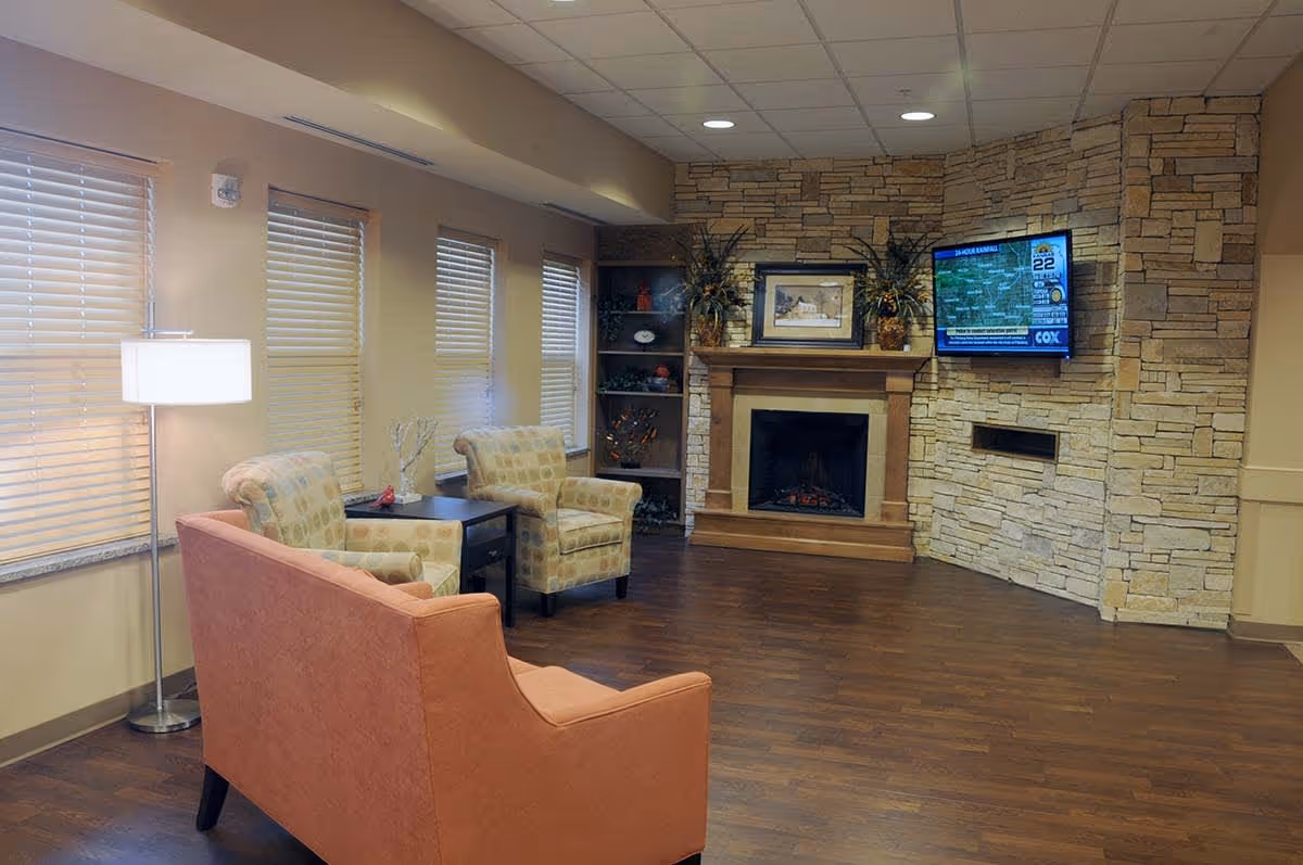 A cozy living room area with a stone fireplace, a mounted flat-screen TV displaying a weather forecast, two patterned armchairs, an orange sofa, a floor lamp, and a small table between the chairs. The room has wooden flooring and several windows with blinds.