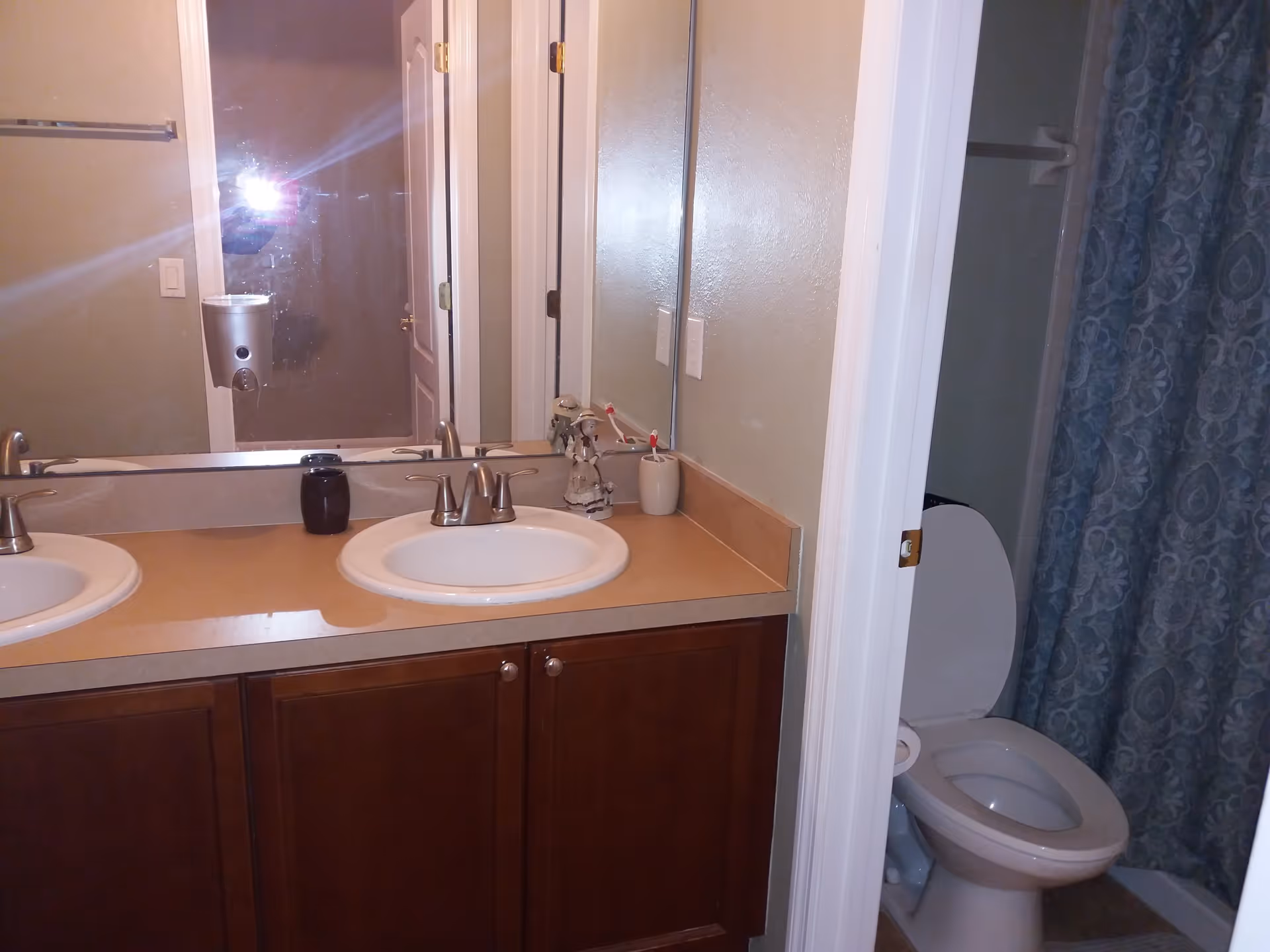 Bathroom with a double sink vanity featuring two faucets and a large mirror above. To the right, there is a toilet with the lid open and a shower area with a blue patterned curtain.