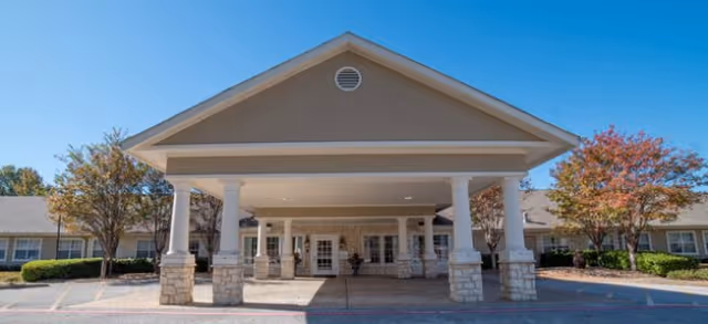 Front exterior view of Brookdale Tyler South facility featuring a covered entrance with white columns and stone bases, surrounded by trees with autumn foliage under a clear blue sky.