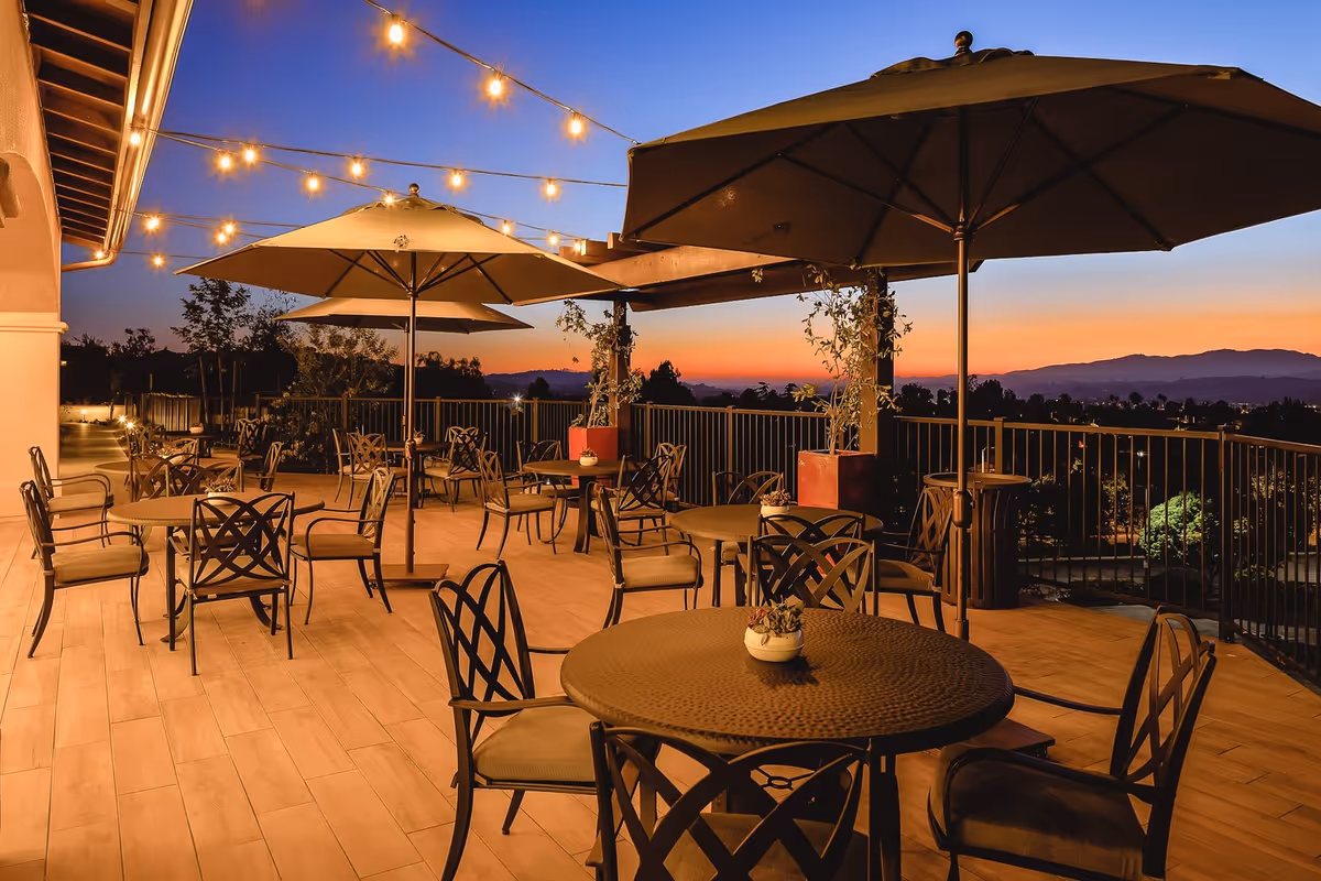 Outdoor patio area at sunset with round tables and cushioned chairs under large umbrellas. String lights are hung overhead, and there are potted plants along the railing. The sky shows a gradient from orange near the horizon to deep blue higher up, with distant mountains visible.