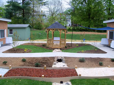 A landscaped courtyard featuring a central wooden gazebo, paved walkways, garden beds, and surrounding single-story buildings.