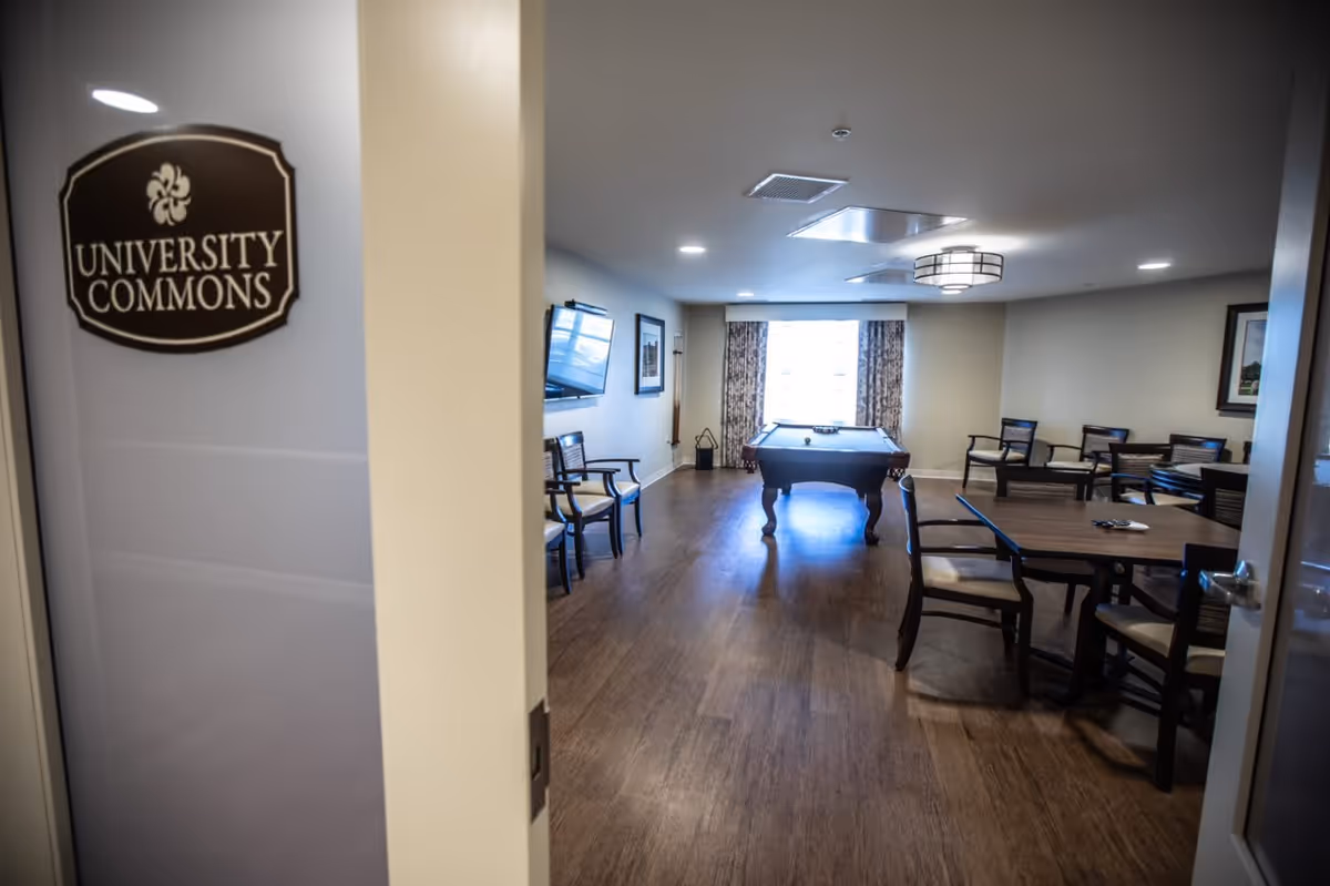 Entrance into a community common room with a pool table, multiple seating tables and chairs, and a TV visible through an open door labeled "University Commons".