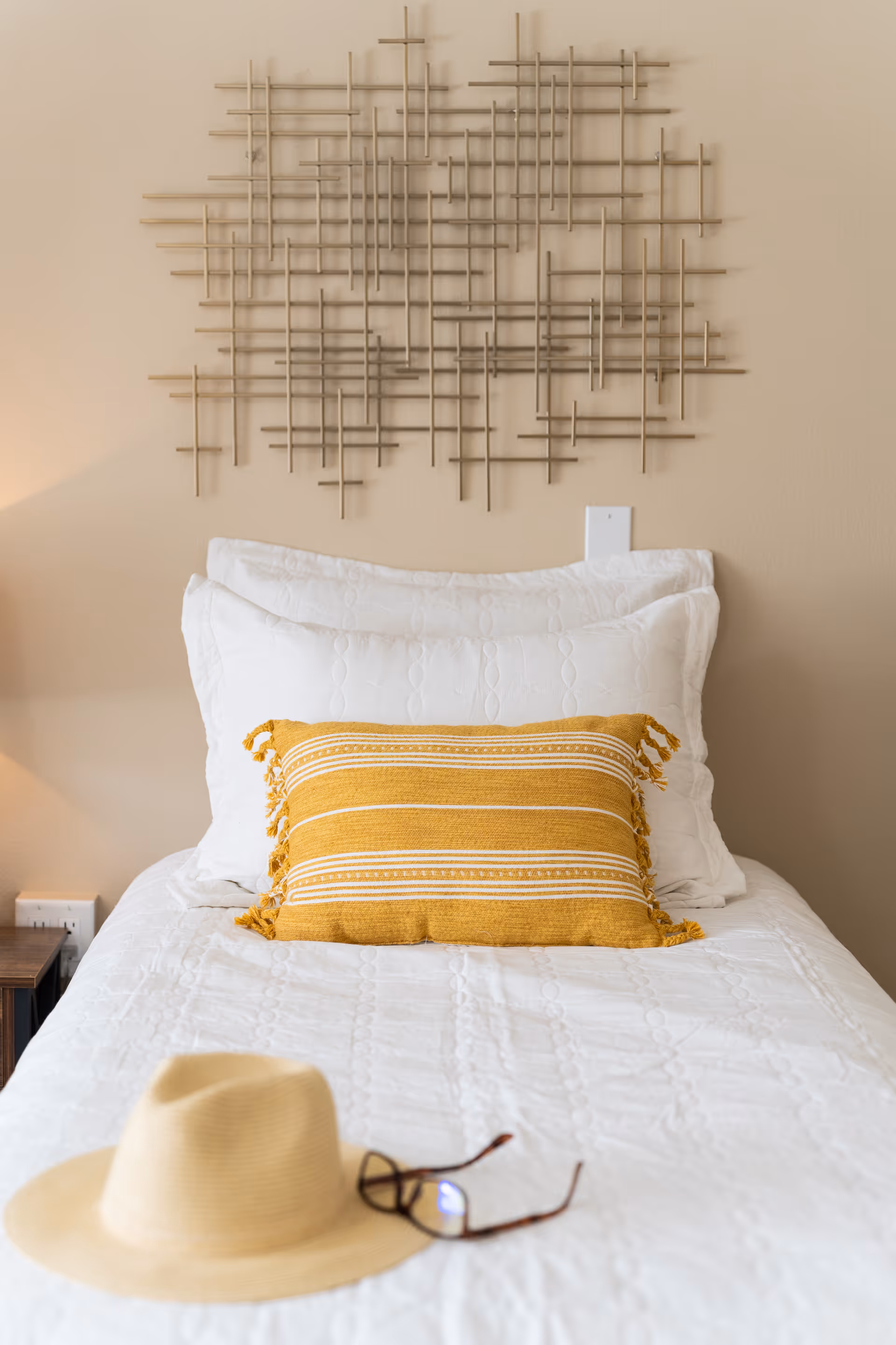 A neatly made single bed with white bedding and a yellow striped decorative pillow. Above the bed is a modern metal wall art piece with intersecting rods. On the bed, there is a beige hat and a pair of eyeglasses. The wall behind the bed is beige.