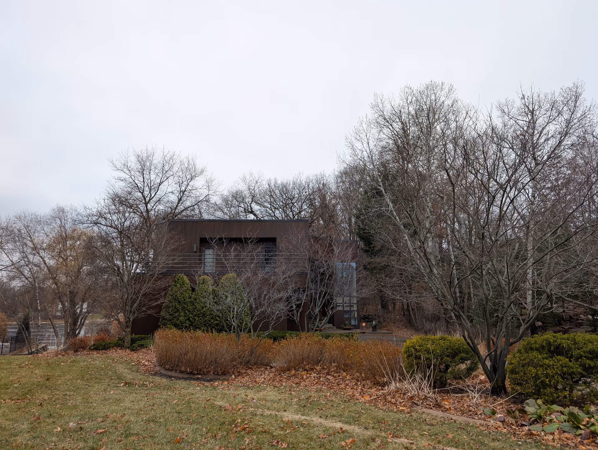 A modern, dark brown building partially obscured by leafless trees and shrubs in a landscaped yard during late autumn or early winter. The sky is overcast and the grass is a mix of green and fallen leaves.