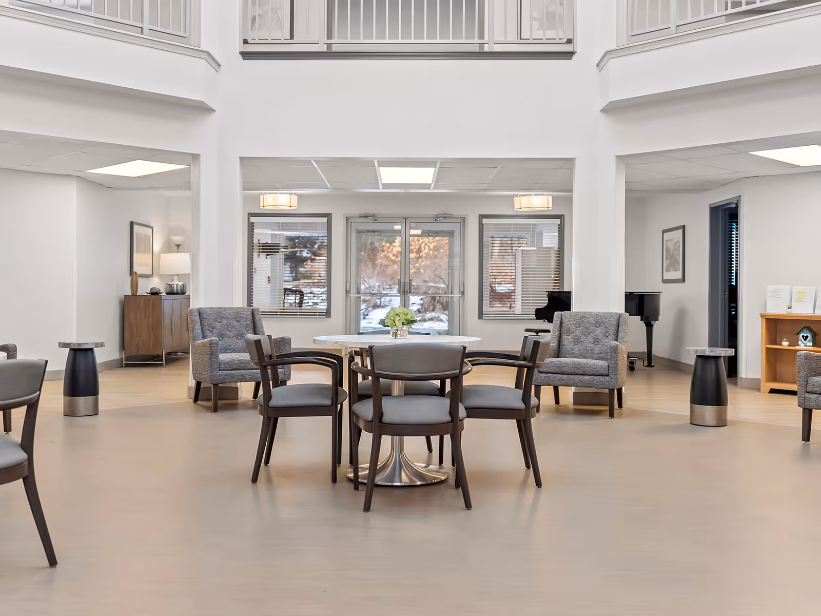 A spacious and bright common area in a senior living facility with modern furniture including a round table with four chairs in the center, two gray armchairs on either side, and a piano in the background near the windows and double glass doors.