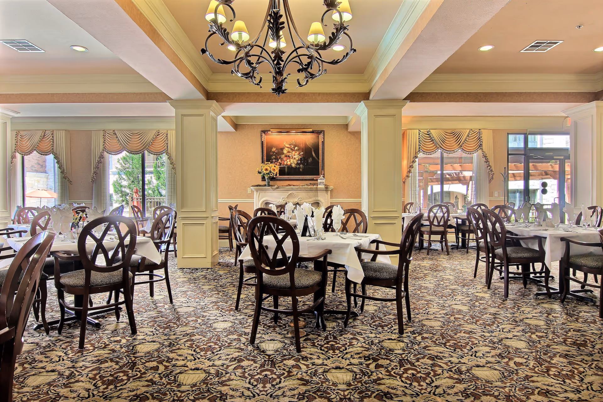 A spacious dining room with multiple round tables covered with white tablecloths and set with glassware and napkins. The room features patterned carpet, large windows with decorative curtains, a chandelier hanging from the ceiling, and a framed painting above a fireplace.