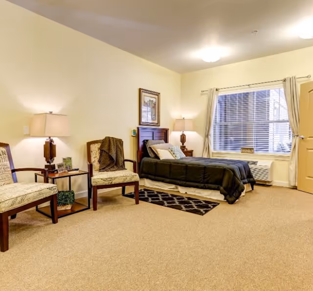 A well-lit bedroom in a memory care community featuring a single bed with dark bedding, two upholstered chairs with a small table between them, two lamps, a framed picture on the wall, a window with curtains, and a beige carpeted floor.