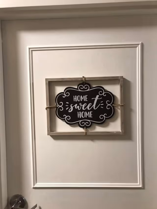 A white door with a decorative sign hanging on it that reads 'Home sweet home' in white cursive and block letters on a black background framed by a rustic wooden frame.