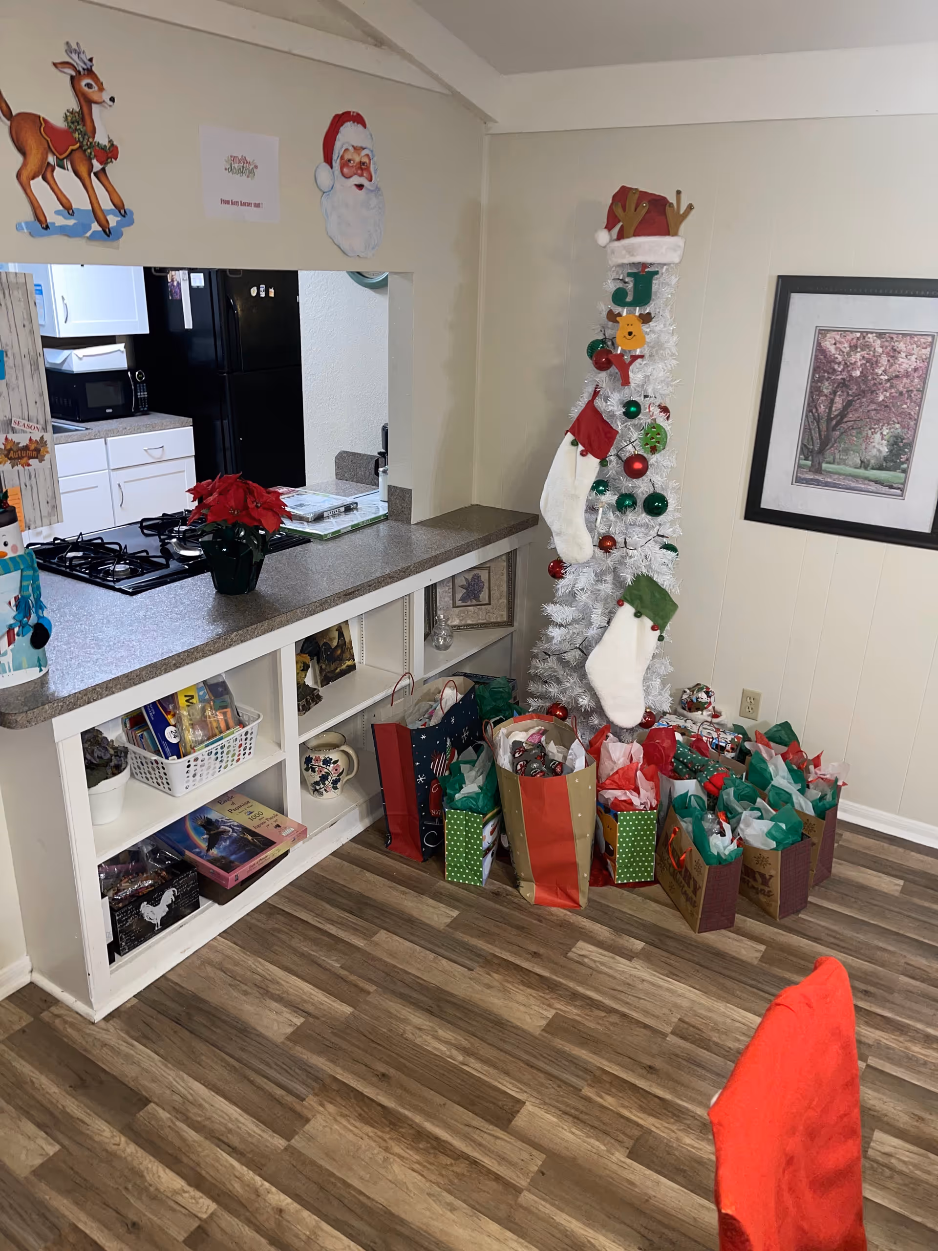 A festive interior corner with a small white Christmas tree adorned with stockings and surrounded by gift bags next to a kitchen pass-through counter.