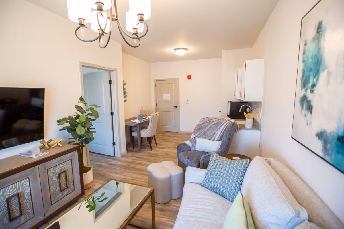 A bright and cozy living area in a senior living facility featuring a beige sofa with green and blue pillows, a gray armchair with a throw blanket, a small ottoman, a glass coffee table, a wooden TV stand with a flat-screen TV mounted above it, a potted plant, and a dining table with chairs in the background. The room has light wood flooring, white walls, a modern chandelier, and a large abstract painting on the wall.