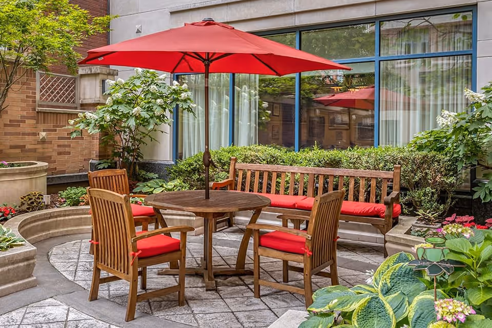 Outdoor patio area with a wooden table and four wooden chairs with red cushions, and a matching wooden bench with a red cushion. A large red umbrella is centered over the table. The patio is surrounded by greenery, plants, and flowers, with a building wall and windows in the background.